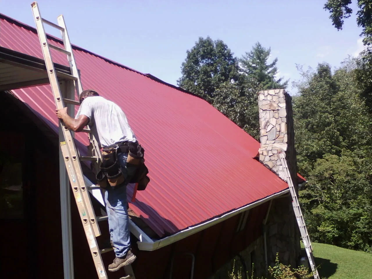 Roofer on a ladder beside a red metal roof and stone chimney — Soffit & Fascia Repair by Roofing Brothers in Clyde